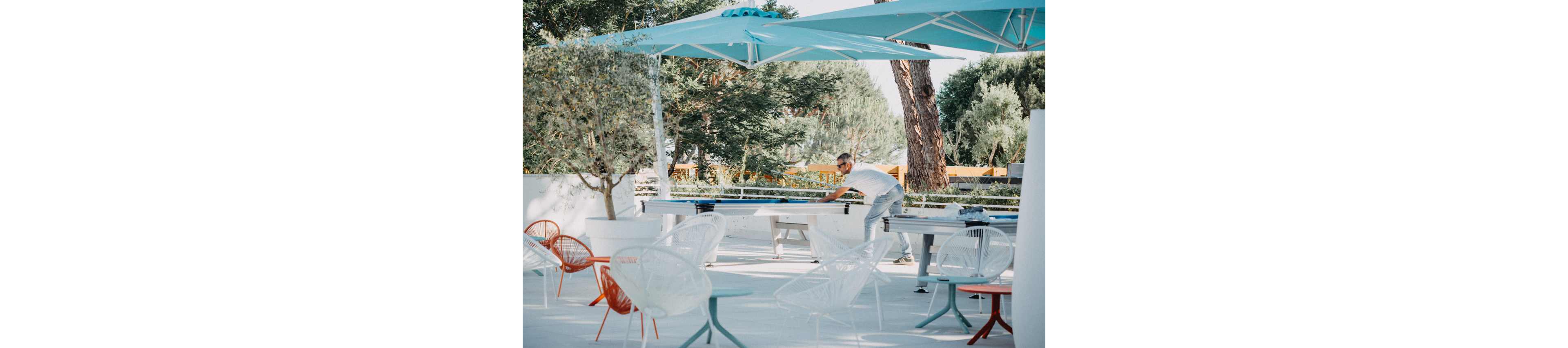 A man leans over an outdoor shuffleboard table in a bright, modern patio area. The setting features white flooring, colorful designer chairs, and large turquoise shade canopies. Trees and greenery surround the space, creating a relaxed, resort-like atmosphere. The overall scene feels casual and summery, with soft natural light and airy decor.