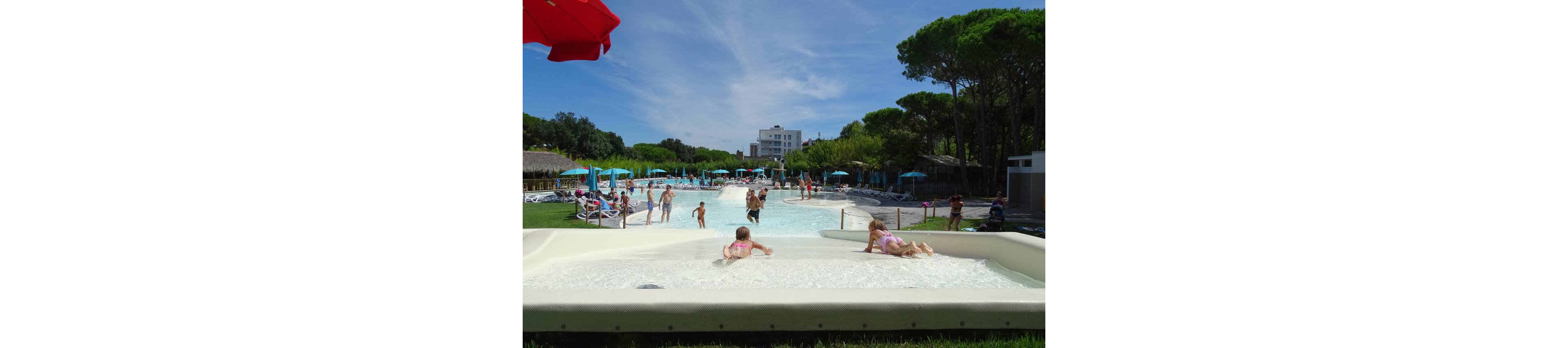 Children play in a shallow wading area at the front of a large outdoor swimming pool. Families and other visitors enjoy the water and sunbathing on loungers under umbrellas in the background. Tall green trees surround the pool complex, with a white building visible in the distance. The scene suggests a relaxed summer holiday atmosphere at a family-friendly resort.