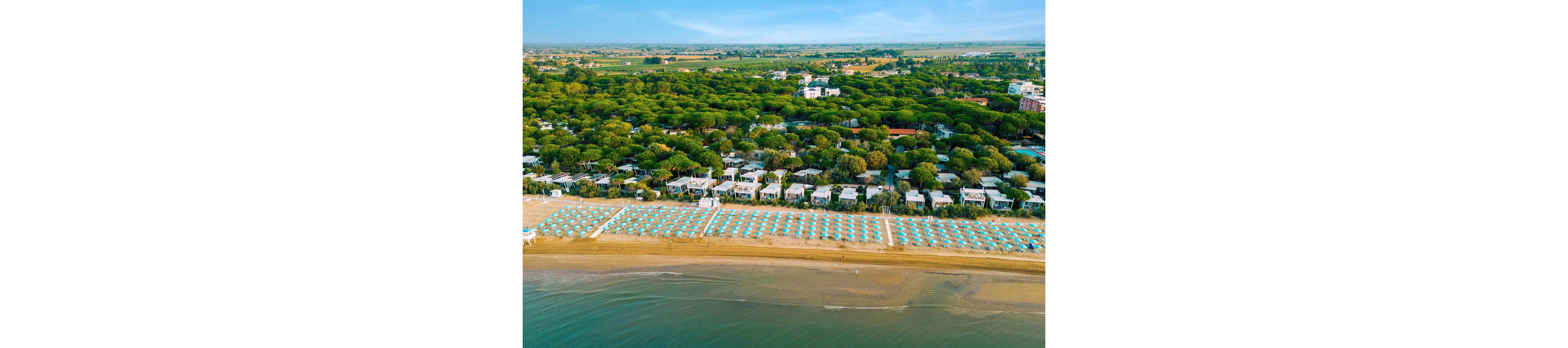 A wide sandy beach lined with rows of turquoise umbrellas stretches along a calm shoreline. Behind the beach, a resort area with small white bungalows is nestled among dense green trees. The scene is captured from above, showing the clear separation between sea, sand, and forested holiday village. The overall atmosphere suggests a peaceful coastal vacation destination on a sunny day.