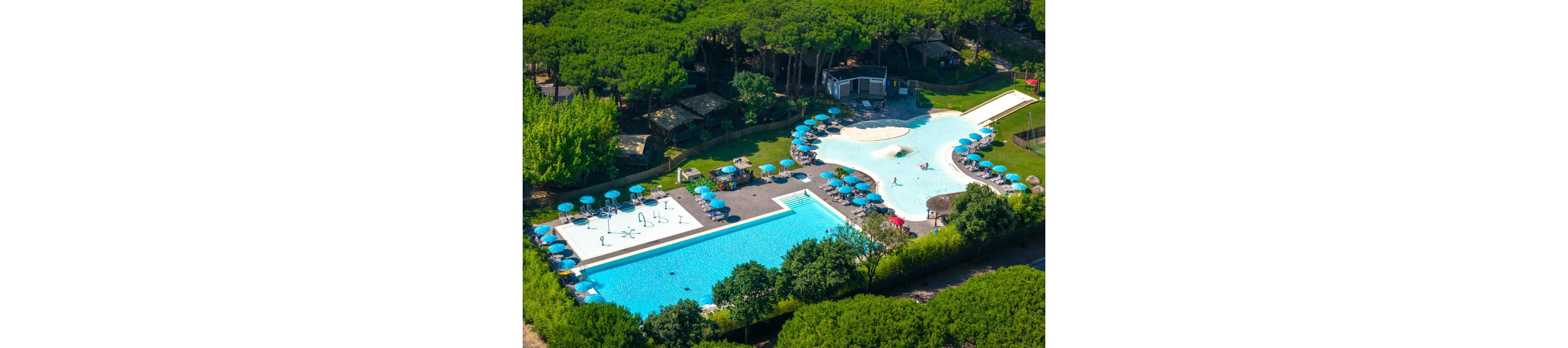 A large outdoor swimming pool complex is surrounded by lush green trees and vegetation. Multiple pools of different shapes include a rectangular lap pool and a lagoon-style pool with shallow areas and a small slide. Blue sun umbrellas and lounge chairs are neatly arranged around the water, with a few people enjoying the facilities. Nearby buildings and pathways suggest a holiday resort or campground setting nestled in a forested area.