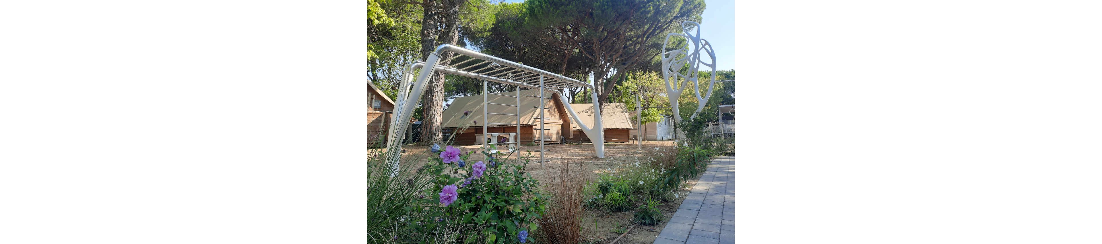 A contemporary white metal climbing structure stands in an outdoor play area surrounded by trees. Behind it are canvas-style cabins or tents, suggesting a campsite or holiday park setting. Flowering plants and ornamental grasses line the foreground next to a paved walkway. A sculptural metal element on the right adds an artistic touch to the recreational space. The overall scene feels calm and family-friendly in natural daylight.