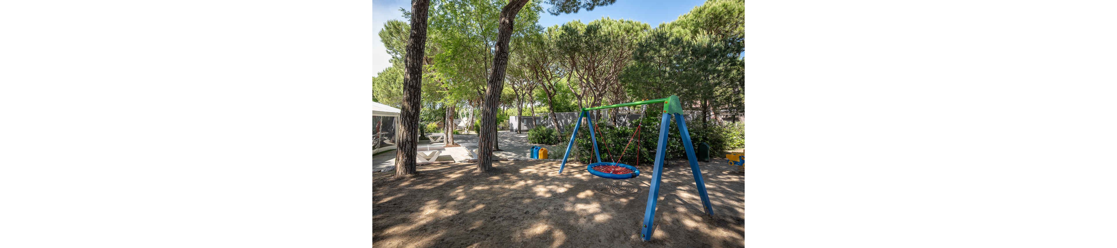 A colorful circular swing hangs from a sturdy metal frame in a shaded outdoor playground. Tall pine trees surround the area, casting dappled light on the sandy ground. In the background, there are lounge chairs, a tent-like structure, and a few plastic play toys. The scene suggests a quiet, family-friendly campsite or park setting on a sunny day.