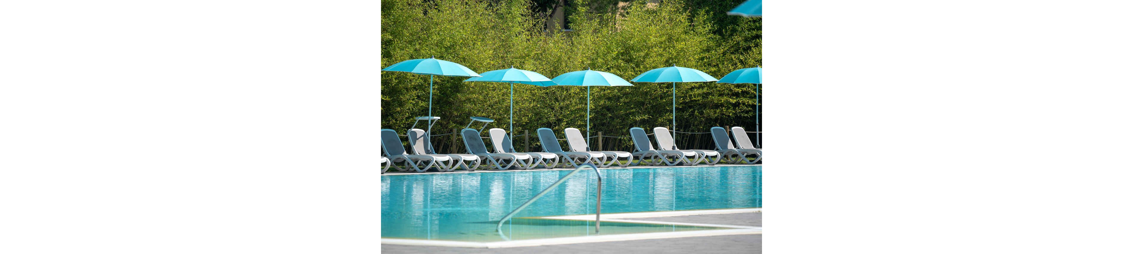 A clear blue swimming pool sits in front of a row of empty lounge chairs. Turquoise umbrellas line the poolside, creating a uniform and relaxing resort setting. The water is calm and reflective, with a metal handrail leading into the pool. Dense green foliage in the background adds privacy and a natural atmosphere.