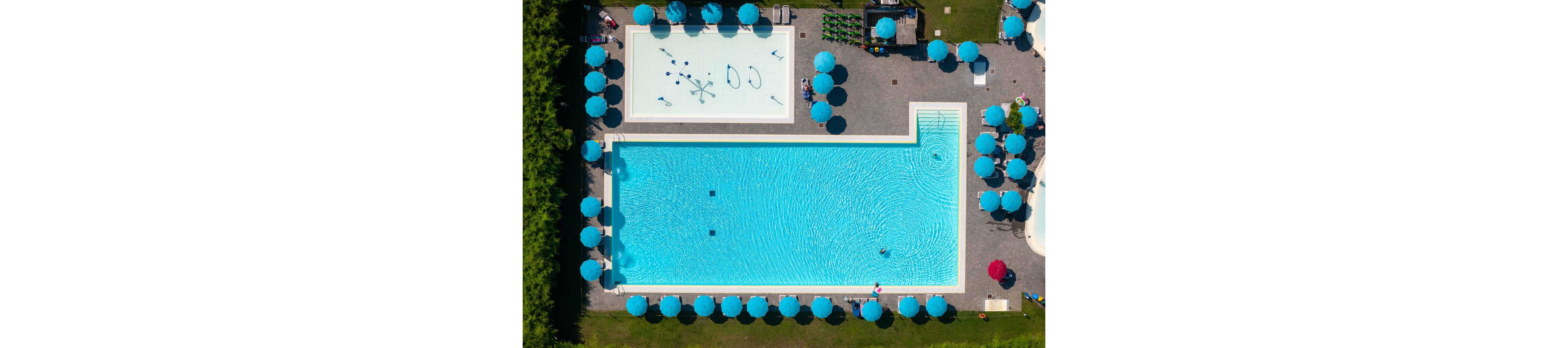 A large rectangular swimming pool and a smaller shallow pool are seen from above in an outdoor recreation area. Bright turquoise water contrasts with the surrounding gray deck and green grass. Numerous blue umbrellas and sun loungers line the perimeter, with a few people visible in and around the water. The scene suggests a sunny, relaxing day at a resort or public pool complex.