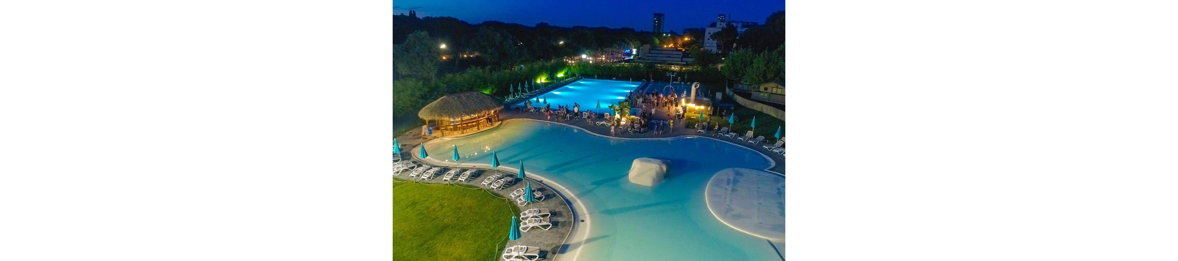 A large outdoor swimming pool complex is shown at night, brightly lit against the dark sky. People gather near a rectangular pool area for a party or event, while nearby lounge chairs and umbrellas sit mostly empty. A thatched-roof bar stands beside the curving shallow pool, adding a tropical touch. Surrounding trees and distant buildings frame the resort, emphasizing a lively yet relaxed vacation atmosphere.