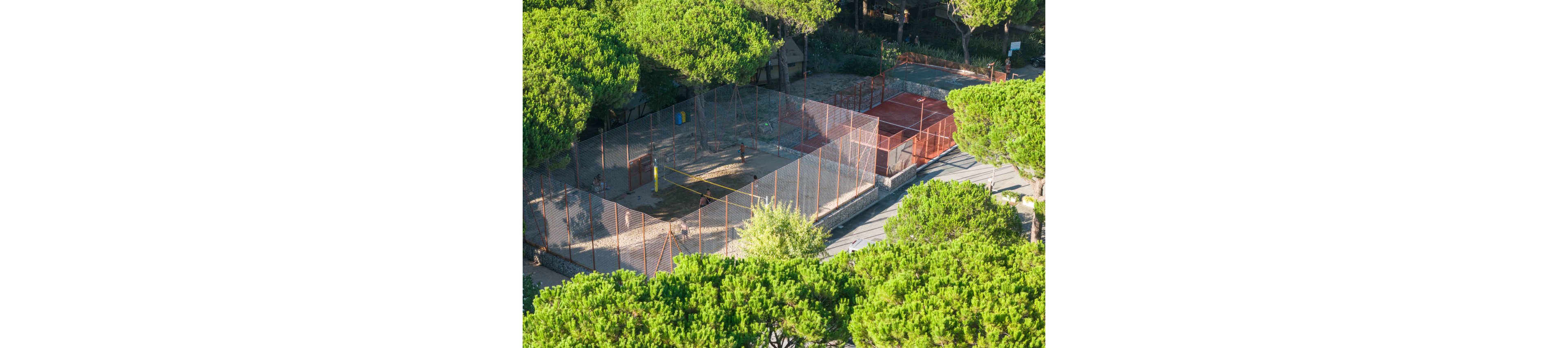 Aerial view of an outdoor sports area surrounded by tall green trees. The scene shows fenced courts, including a sand volleyball court with a yellow net and a hard-surface court beside it. Orange fencing and pathways define the recreational zone within the wooded setting. The image conveys a sunny, peaceful atmosphere in a park or resort environment.
