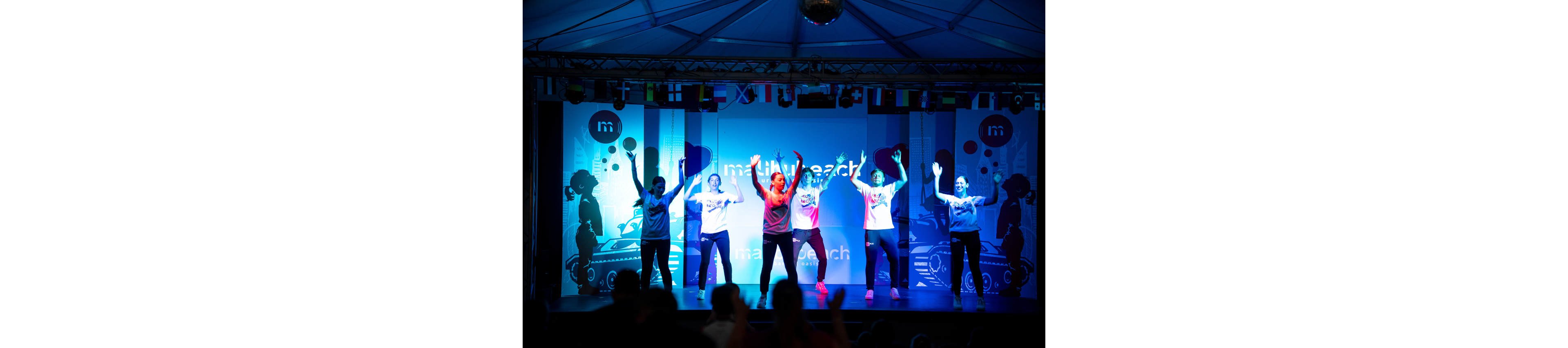 A group of performers dances energetically on a brightly lit stage. They are wearing casual outfits and are positioned in a line with their arms raised in unison. The background features colorful blue and purple lighting with graphic silhouettes and text panels. Audience members are visible as dark silhouettes in the foreground, emphasizing the concert-like atmosphere.
