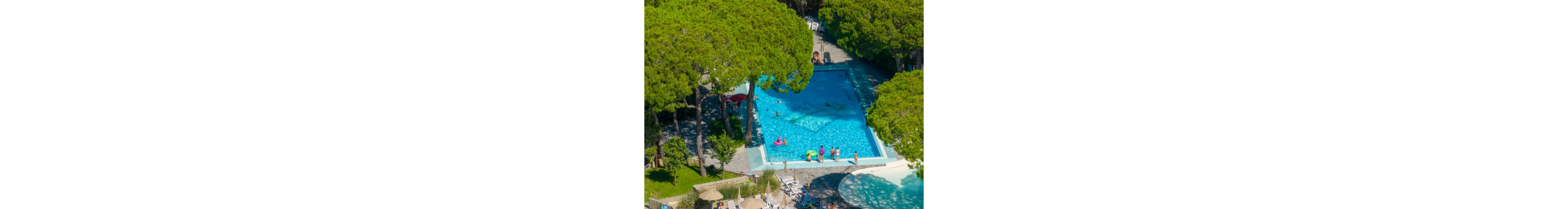 An outdoor swimming pool is surrounded by tall green trees in a forested resort setting. Several people are in and around the bright blue water, some swimming and others standing at the pool edge. Sun loungers and umbrellas are arranged nearby, suggesting a relaxing vacation atmosphere. The scene is captured from above, emphasizing the rectangular pool and the contrast between turquoise water and lush greenery.