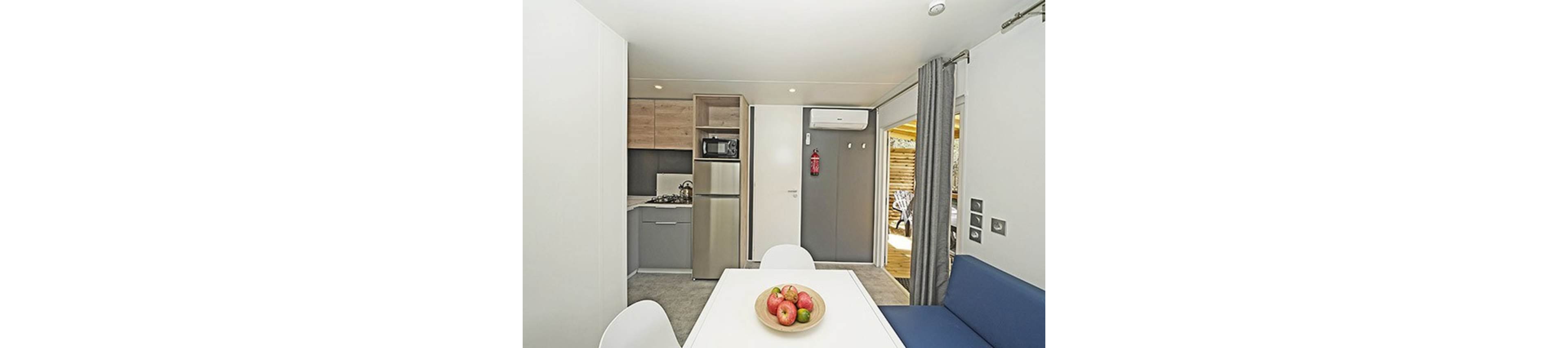 Bright compact studio interior featuring a small kitchen and dining table. The space includes built-in cabinets, a stovetop, sink, microwave, and tall refrigerator. A white table with a bowl of apples sits beside a blue bench and white chairs. Neutral walls, grey flooring, and a sliding door with curtains open to an outdoor patio area.