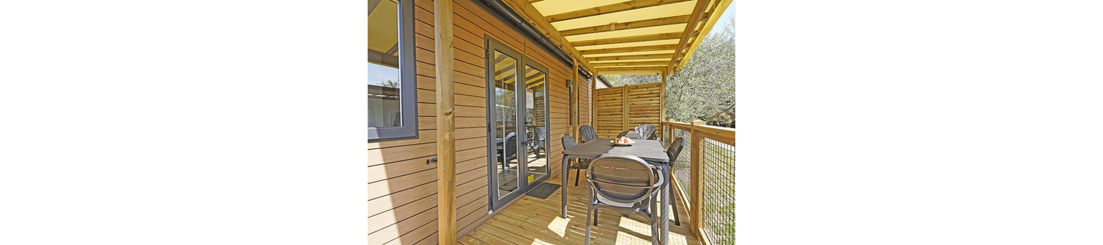 A covered wooden terrace features a dark outdoor dining table with several matching chairs. The space is attached to a modern cabin with horizontal wood siding and glass doors. A bowl of fruit sits on the table, adding a small accent of color. The terrace is partially enclosed with wooden slats and a railing, overlooking surrounding greenery.