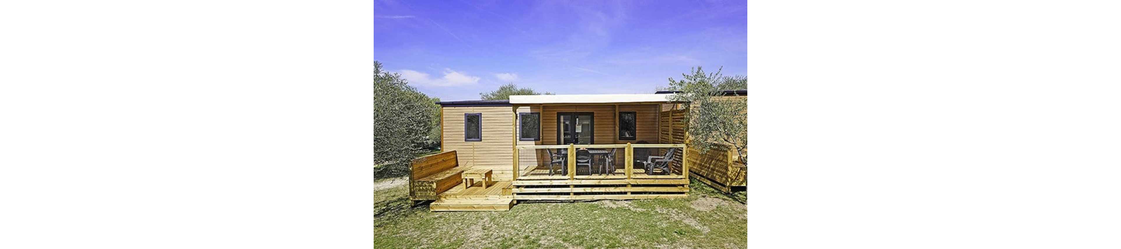 A contemporary wooden mobile home stands in an open outdoor setting under a clear blue sky. The structure features a covered front terrace with outdoor chairs and a table. A built-in wooden bench and table sit on an adjacent deck area, emphasizing space for relaxation and socializing. Surrounding trees and sparse grass suggest a natural, campground-like environment ideal for vacations.