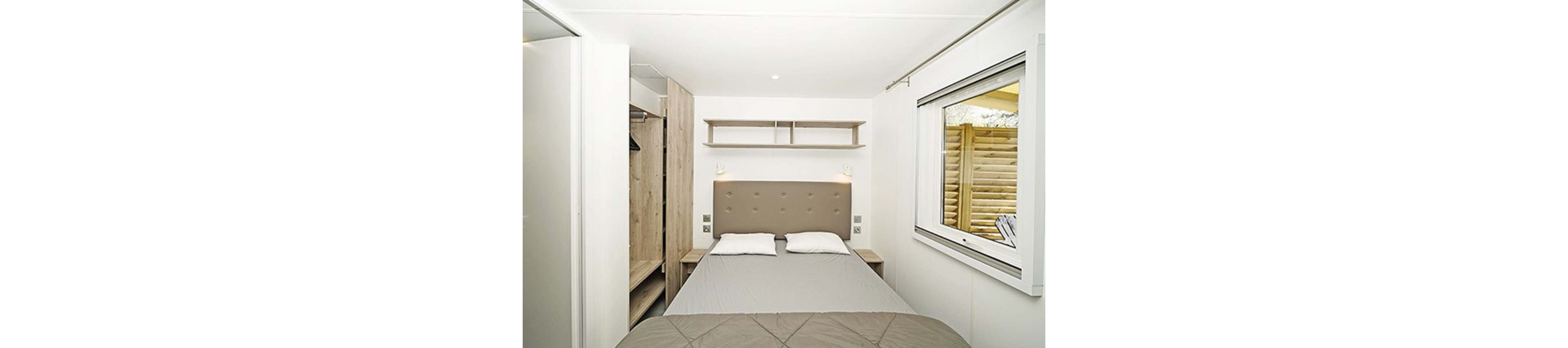 A small modern bedroom features a double bed with a padded headboard and two pillows. Built-in wooden wardrobes and open shelving provide storage on the left side of the room. A large window on the right lets in natural light and offers a view of an exterior wooden screen. The color palette is neutral with white walls and beige and gray textiles, creating a clean and minimalist atmosphere.