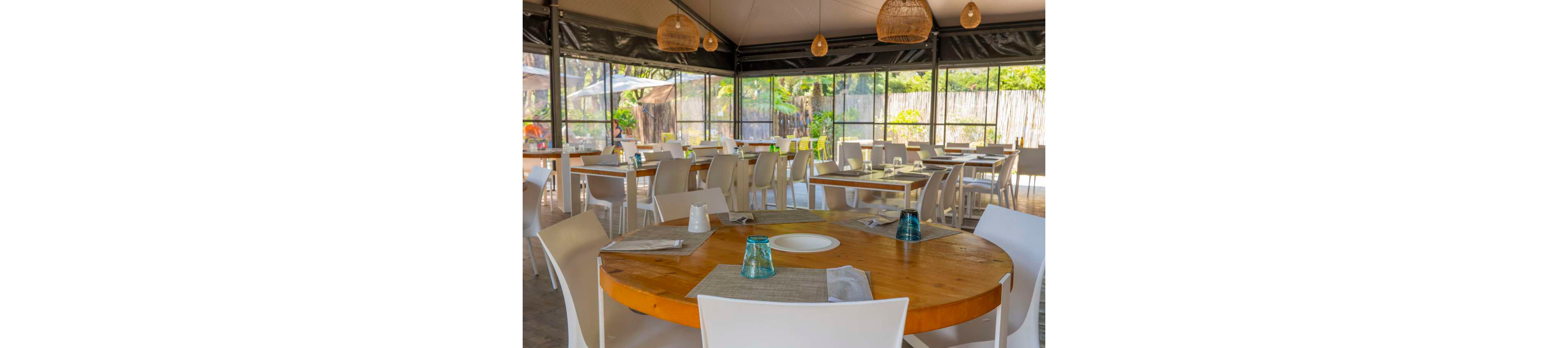 A bright restaurant dining area features round wooden tables set with placemats, plates, and upside-down glasses. White modern chairs surround the tables, creating a clean and contemporary look. Large windows and open sides reveal greenery and a bamboo fence outside, suggesting an outdoor or semi-open setting. Warm hanging lamps and natural light contribute to a relaxed, inviting atmosphere.