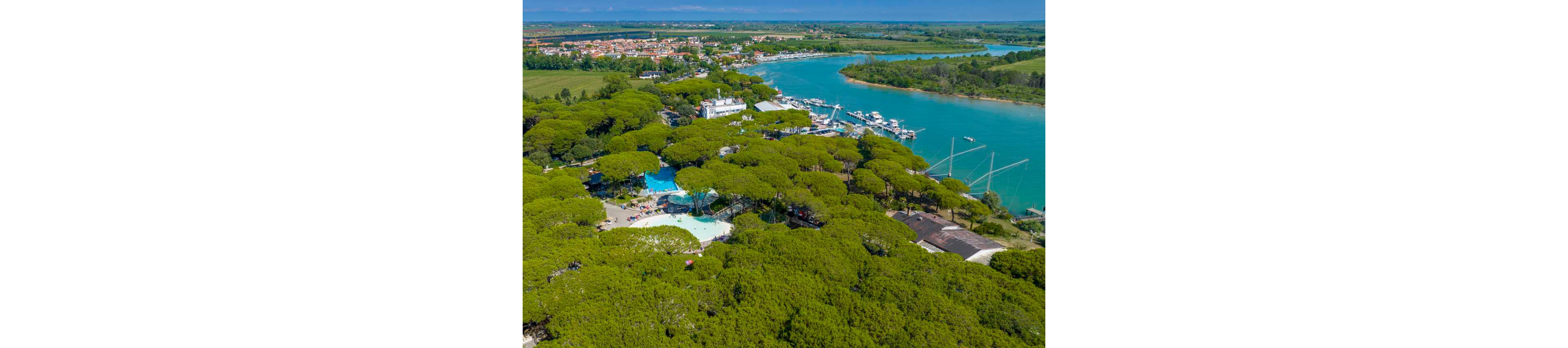 A high-angle aerial view shows a lush green resort area surrounded by dense trees. A bright turquoise river curves alongside a marina filled with docked boats and yachts. Within the trees, swimming pools and recreational areas are visible, suggesting a holiday or camping village. In the distance, a small town and open countryside stretch toward the horizon under a clear blue sky.