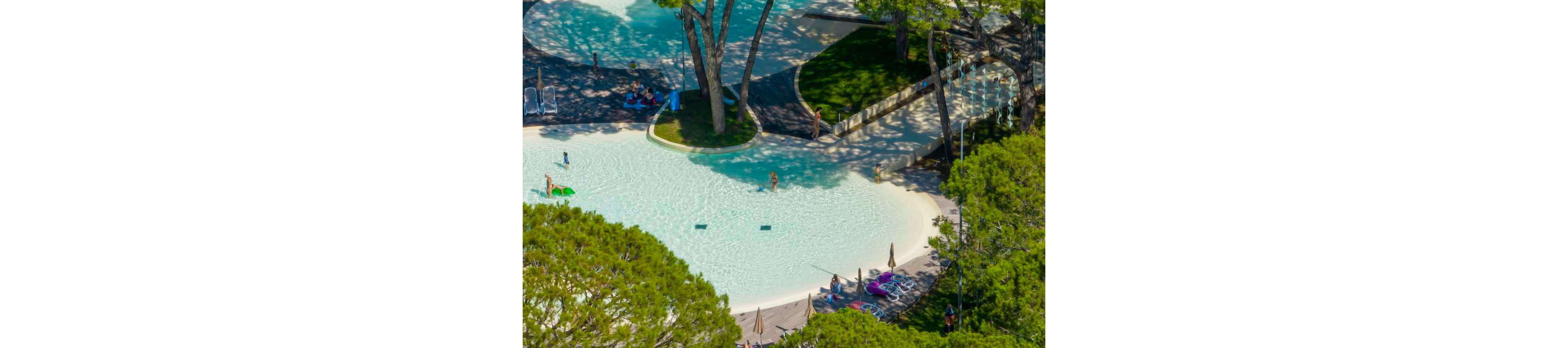 A shallow resort swimming pool is nestled among tall green trees, creating a shaded leisure area. A few people wade and play in the clear water near the gently sloping pool edge. Sun loungers and colorful towels are arranged around the poolside deck. The scene suggests a relaxed holiday atmosphere in a family-friendly outdoor setting.