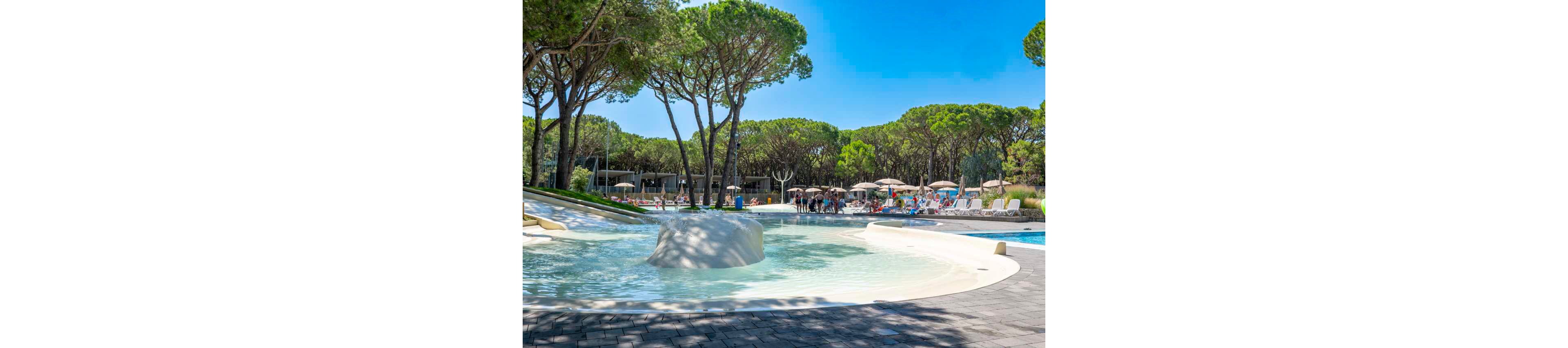 An outdoor resort swimming pool is surrounded by tall pine trees and modern low-rise buildings. Guests relax on sun loungers and under umbrellas along the poolside. The shallow foreground pool features a smooth white island and gently sloping edges. Bright blue sky and clear water create a summery vacation atmosphere.