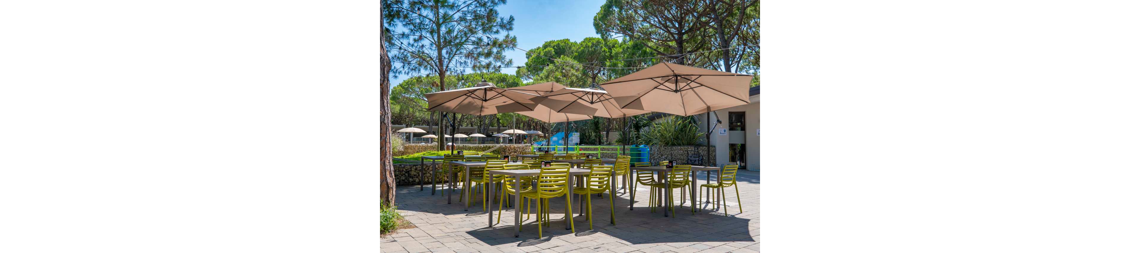 An outdoor café terrace features several tables with bright green chairs arranged on a stone-paved patio. Large beige umbrellas provide shade over the seating area, creating a comfortable place to relax. In the background, lush green trees, hedges, and additional parasols suggest a resort or garden setting. A low stone wall and nearby building complete the peaceful, sunny atmosphere.