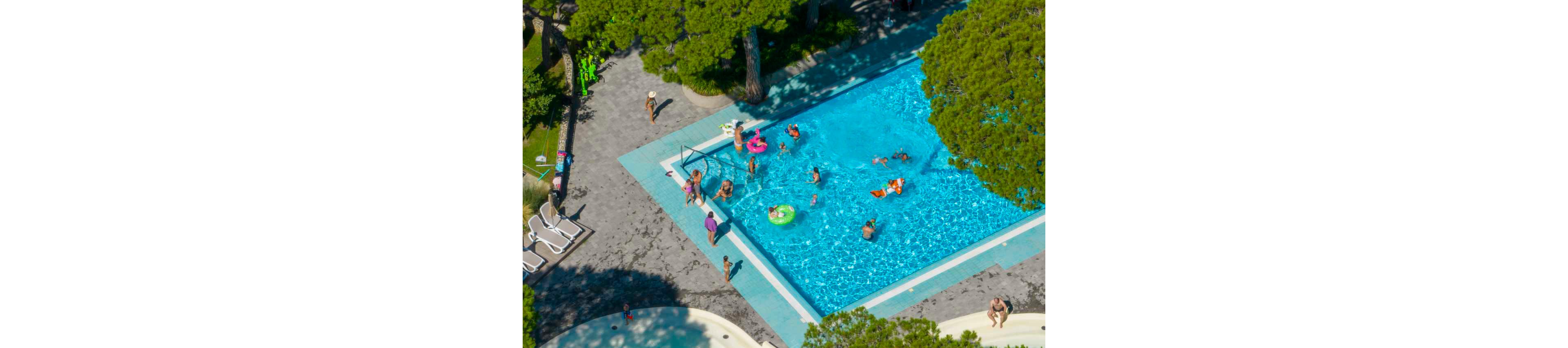 A bright outdoor swimming pool is filled with people enjoying the water. Children and adults play with inflatable rings and toys while others swim or stand in the shallow areas. The pool is surrounded by trees, lounge chairs, and paved walkways, creating a relaxed vacation atmosphere. Sunlight highlights the clear blue water and the vibrant colors of swimwear and floats.