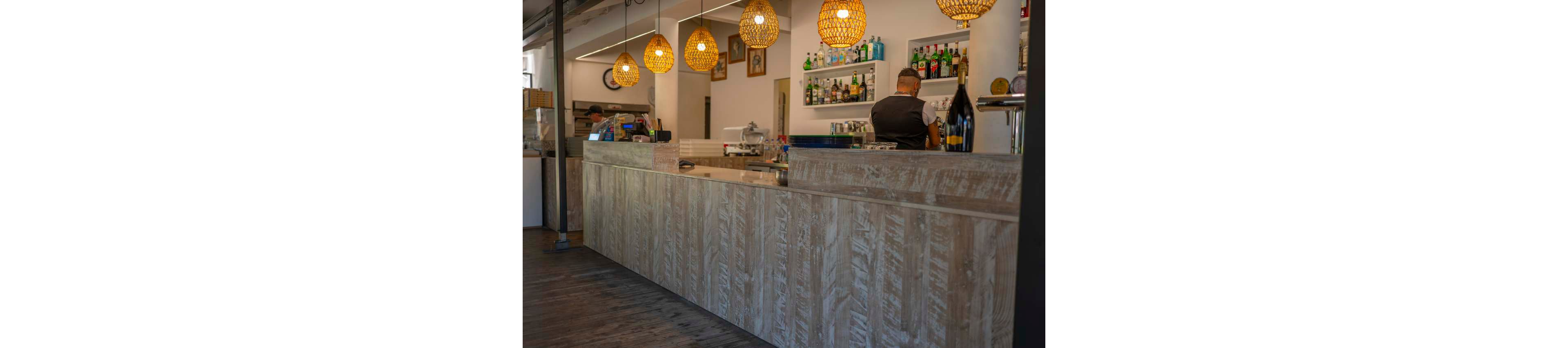 A contemporary bar interior features a long wooden counter with a textured finish. A bartender stands behind the bar preparing drinks, with shelves of assorted bottles and glassware on the wall. Warm woven pendant lights hang from the ceiling, casting a cozy glow over the space. Another staff member works in the background near the kitchen area, adding to the active yet relaxed atmosphere.