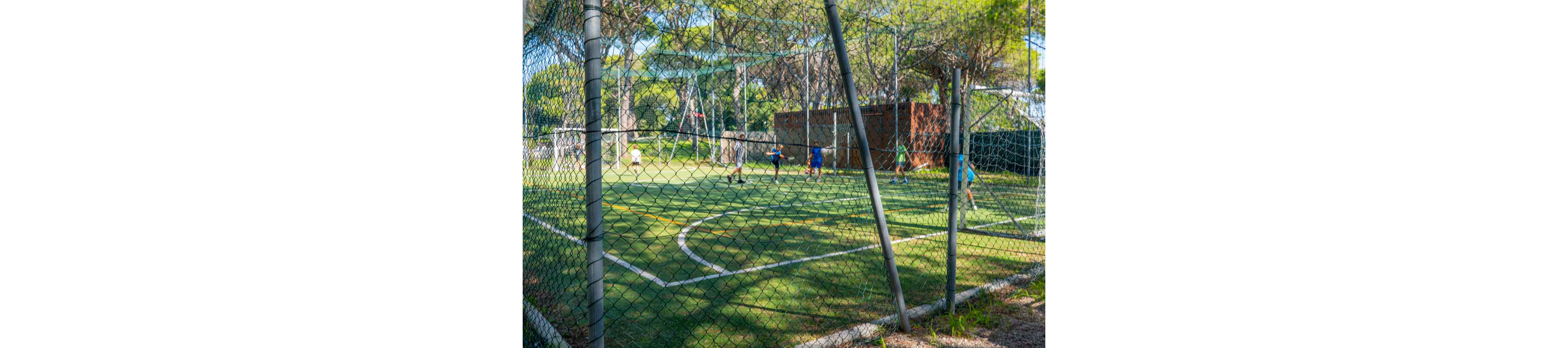 A group of children play soccer on a small outdoor field surrounded by a tall wire fence. The artificial turf pitch is marked with white lines and is set in a green park with many trees. A brick building and netting structures are visible in the background. Sunlight filters through the trees, creating a bright and relaxed recreational atmosphere.