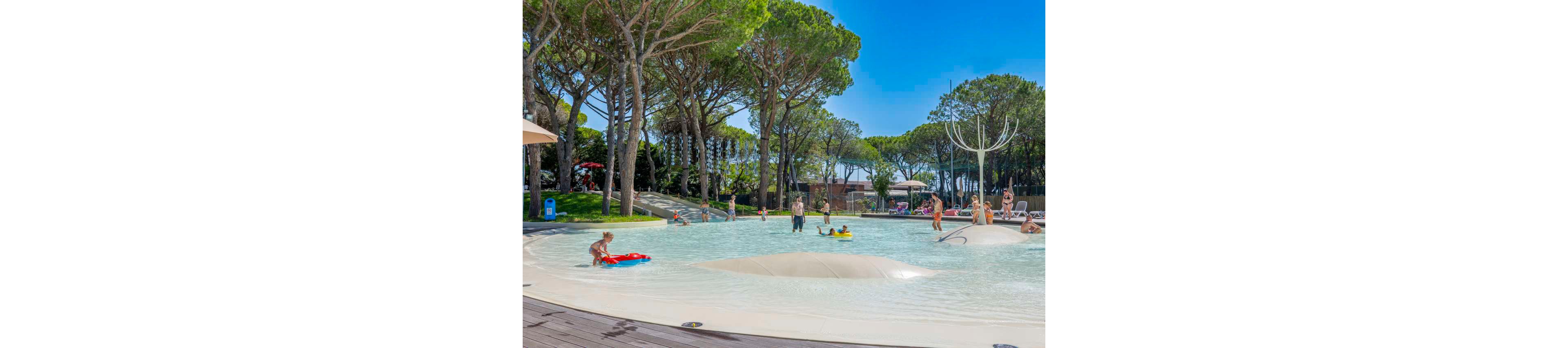 Children and adults enjoy a shallow splash pool surrounded by tall pine trees. The scene shows kids wading, playing with inflatables, and climbing on soft white mounds in the water. Parents and caregivers stand nearby, supervising and relaxing under the clear blue sky. The natural setting and bright daylight create a cheerful, family-friendly atmosphere.
