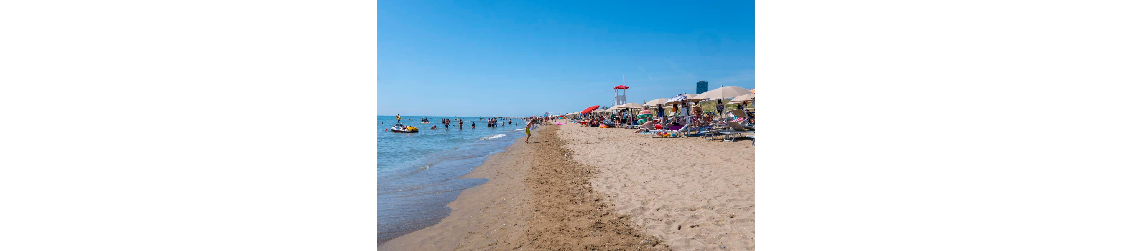 A busy sandy beach is filled with people relaxing on lounge chairs under umbrellas and playing in the shallow sea. Swimmers and children enjoy the calm water near the shoreline, while others walk along the wet sand. Colorful beach umbrellas, inflatables, and a lifeguard tower add visual interest to the sunny scene. The clear blue sky and distant buildings suggest a popular coastal resort on a bright summer day.