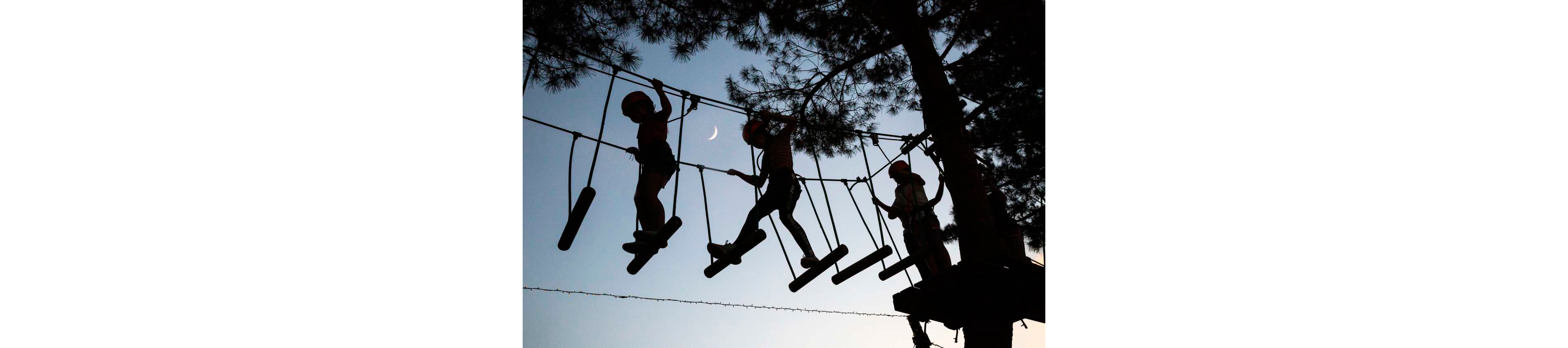 Three children wearing helmets are silhouetted as they navigate a high ropes course suspended between trees. The scene is set outdoors at dusk, with a clear sky and a crescent moon visible in the background. The kids balance on hanging wooden planks while clipped into safety lines. The overall mood is adventurous and playful, emphasizing outdoor recreation and teamwork.