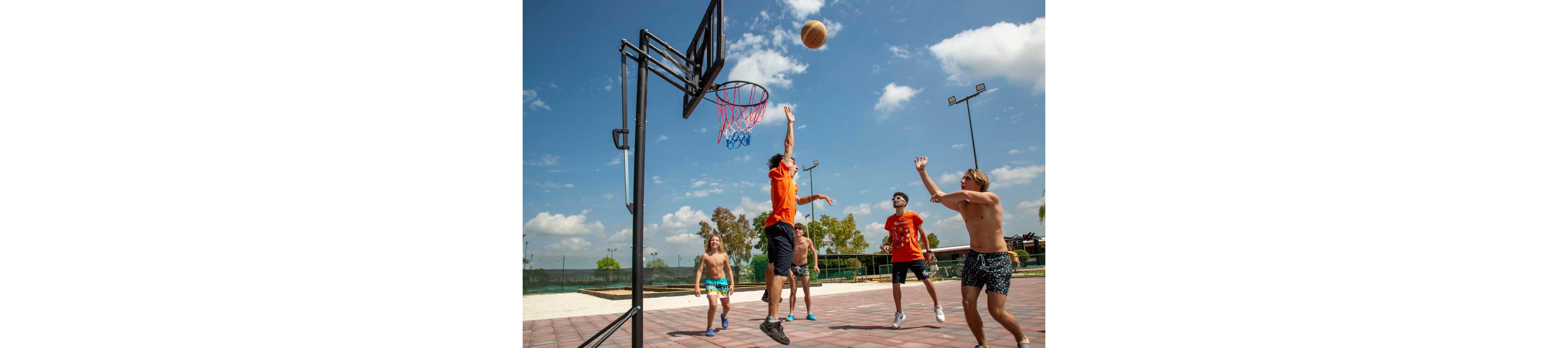 A group of young adults play a casual basketball game on an outdoor court. One player in an orange shirt jumps to shoot the ball toward the hoop while others defend and watch. The scene is set under a bright blue sky with scattered clouds and surrounding greenery. The atmosphere feels energetic, summery, and relaxed, suggesting a fun recreational moment with friends.