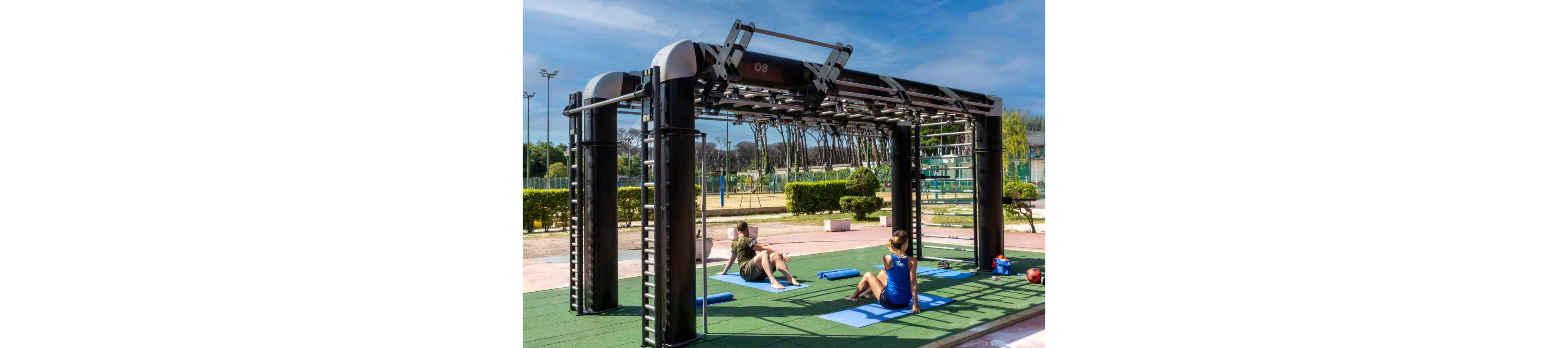 Two people exercise on mats beside a large outdoor fitness rig in a sunny park setting. The black metal structure features multiple bars and ladders for bodyweight training, with the number 08 visible on the top beam. Surrounding the workout area are trees, hedges, and sports courts under a clear blue sky. The scene suggests a relaxed yet active atmosphere focused on health and fitness.