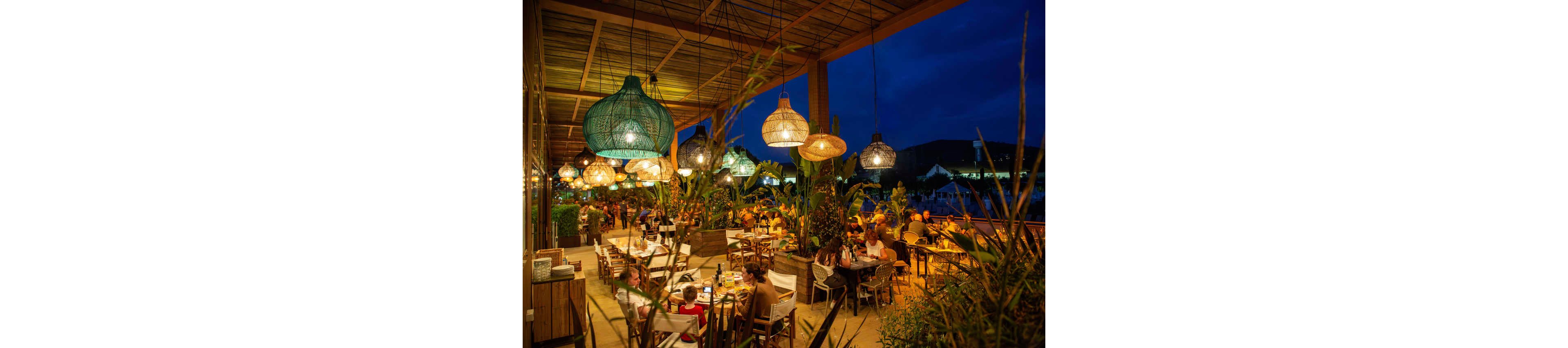 A bustling open-air restaurant terrace is filled with diners seated at wooden tables under a large covered roof. Warm hanging lanterns and woven light fixtures create a cozy glow against the deep blue evening sky. Lush tropical plants surround the seating area, adding a natural, vibrant atmosphere. People of various ages enjoy food and conversation in a relaxed, social setting.