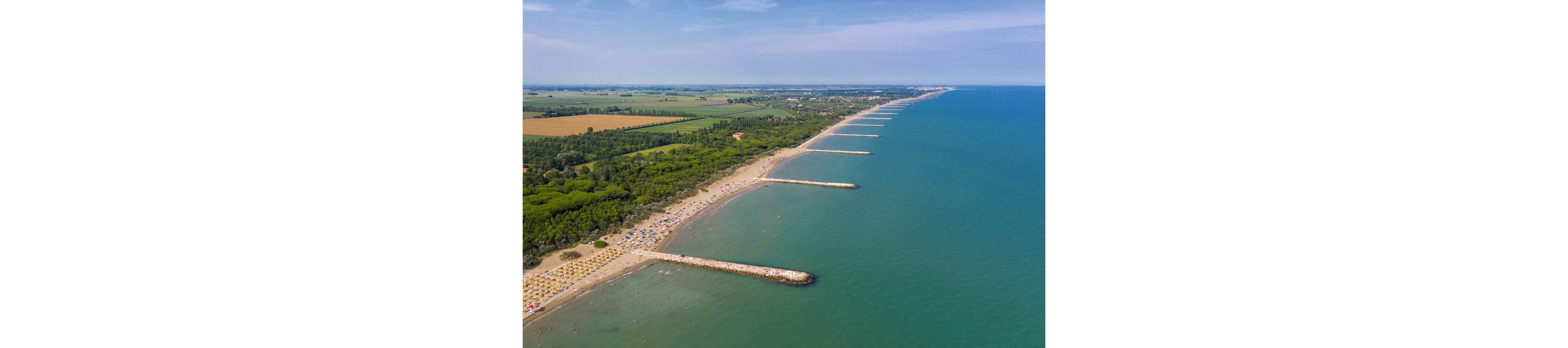 A wide aerial view shows a long sandy beach bordered by calm blue sea. Parallel stone breakwaters extend into the water at regular intervals along the shoreline. Colorful umbrellas and sunbathers line the beach, backed by dense green trees and agricultural fields. The scene suggests a peaceful summer day on a popular coastal destination.