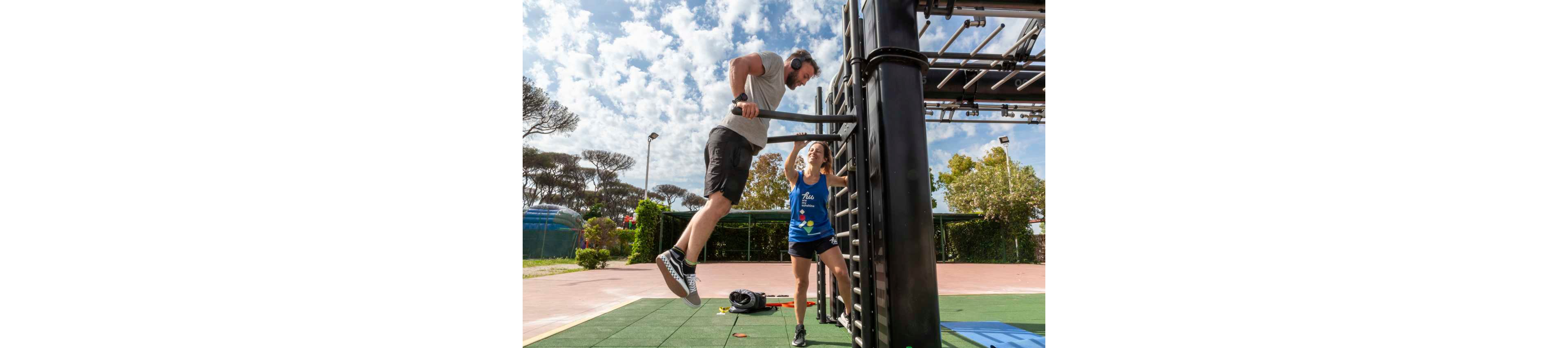 A man performs pull-ups on an outdoor fitness structure while a woman stands nearby, encouraging him. The scene takes place on a sunny day in a park-like setting with trees, greenery, and a clear sky filled with scattered clouds. Exercise equipment and a gym bag lie on the ground, emphasizing a workout environment. The overall mood is energetic and motivational, highlighting functional training and teamwork.