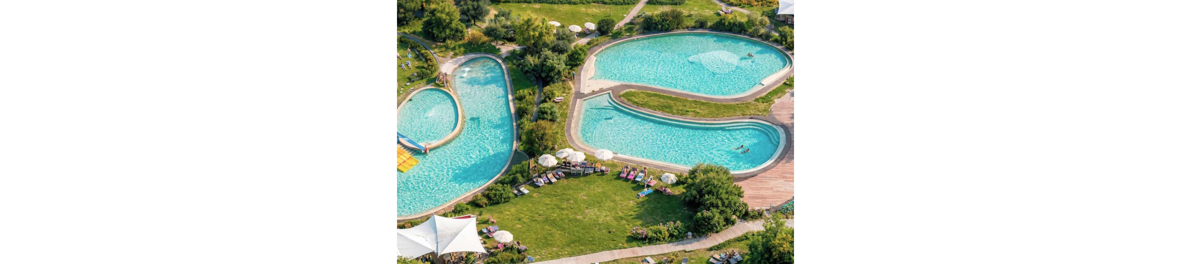 A large outdoor swimming pool complex is seen from above, featuring multiple interconnected pools surrounded by green lawns. Sun loungers and umbrellas are arranged neatly around the water, with a few people relaxing and swimming. Curved wooden walkways and landscaped trees create an inviting resort atmosphere. The scene suggests a warm, sunny day ideal for leisure and vacation.
