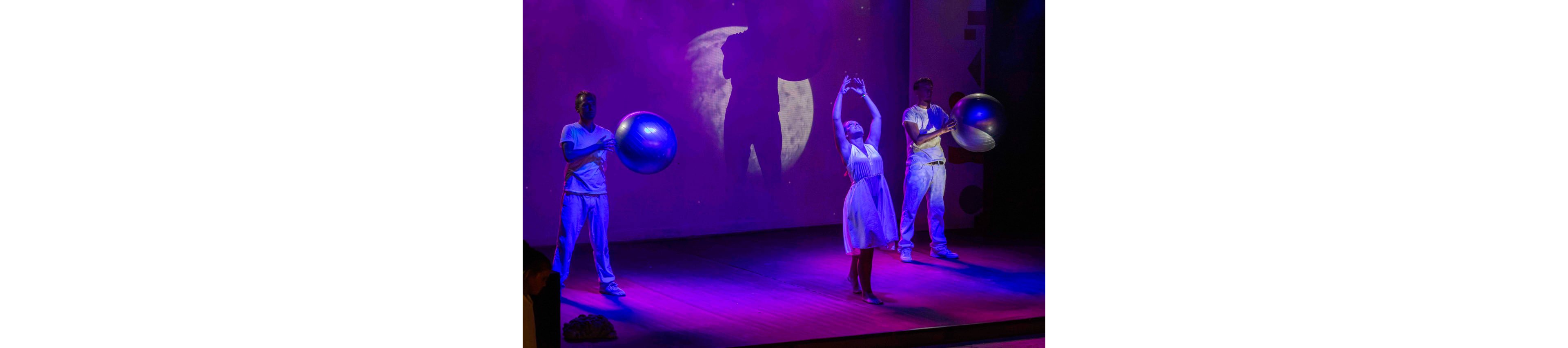 Three performers stand on a stage bathed in purple and blue light. The central figure in a white dress raises their arms gracefully, suggesting a dance or theatrical movement. Two other performers in white outfits hold large exercise balls on either side, adding a modern, abstract element. A projected full moon and shadowy silhouette appear on the backdrop, enhancing the dramatic and surreal atmosphere.