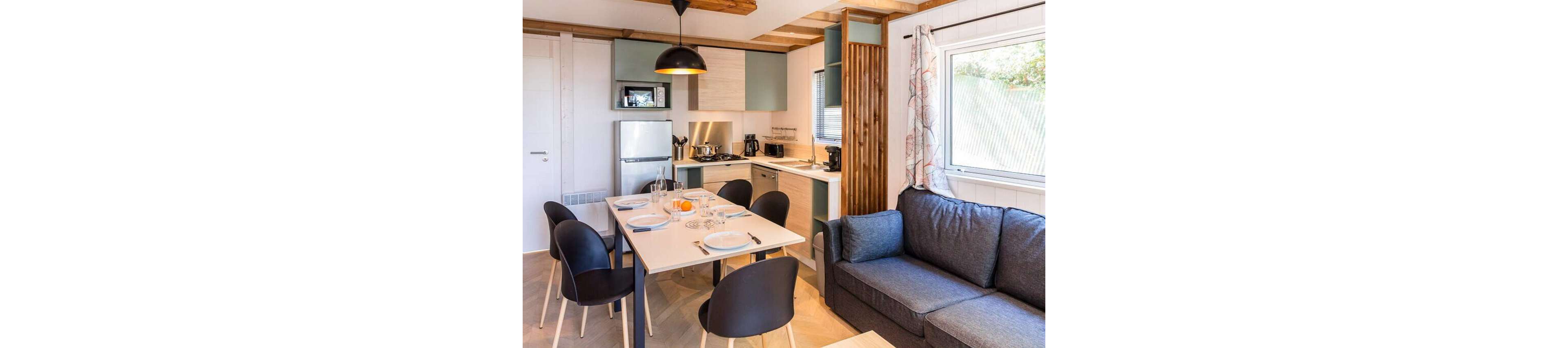 Bright open-plan interior featuring a compact modern kitchen and adjacent dining table. The table is set with plates, glasses, and cutlery, suggesting a meal about to be served. A gray fabric sofa sits beside a large window with patterned curtains, adding comfort to the space. Light wood finishes, soft pastel cabinetry, and black chairs create a contemporary yet inviting atmosphere.