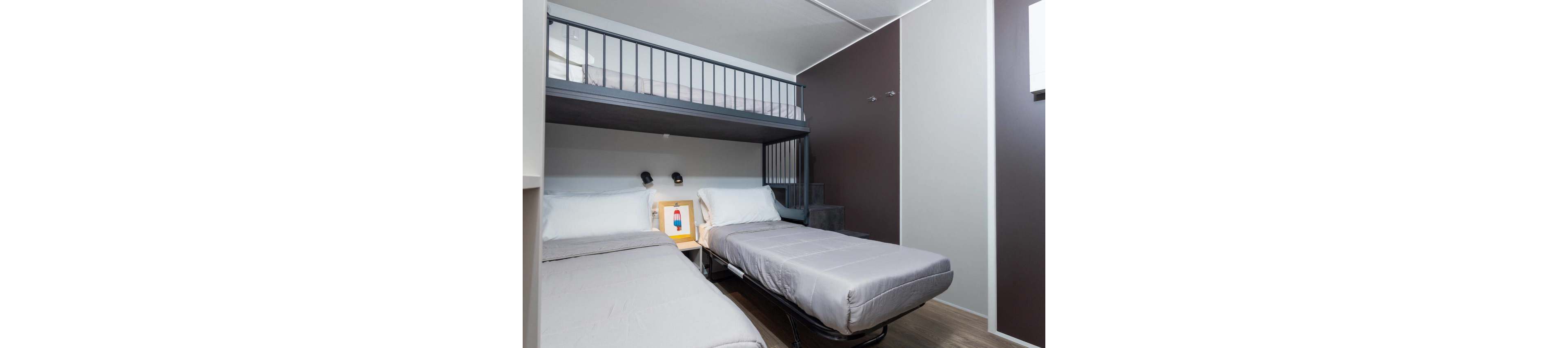 A small contemporary bedroom features a bunk bed above two single beds arranged in an L-shape. The space has clean lines, neutral gray and white walls, and wood-look flooring that emphasizes a minimalist design. Two wall-mounted reading lights are positioned above the pillows, and a framed colorful print sits on the bedside surface. A small staircase leads up to the upper bunk, highlighting the room’s efficient use of vertical space.