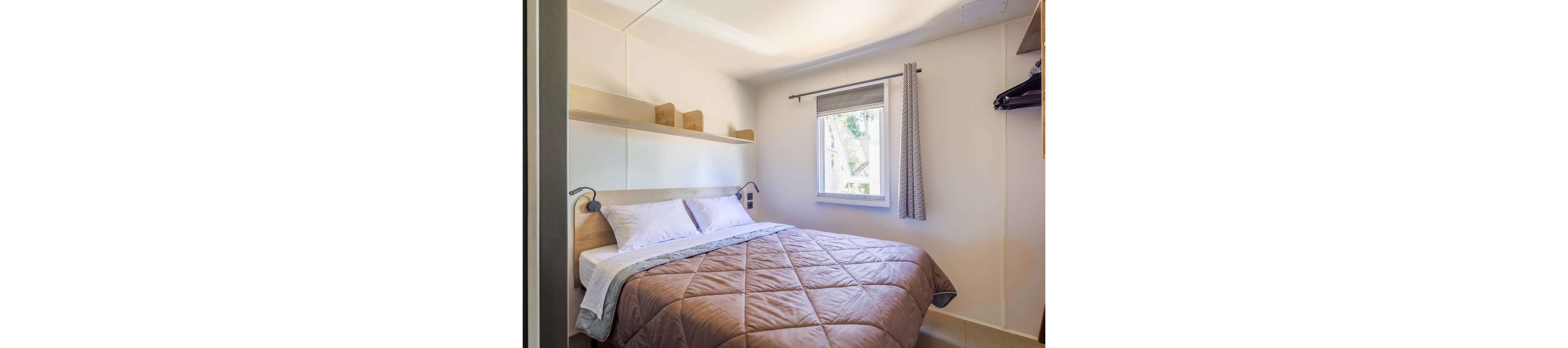 A compact bedroom features a neatly made double bed with a quilted beige comforter and white pillows. The room has light-colored walls, a small window with a gray roller blind and curtain, and built-in shelving above the bed. A clothes rail with hangers is visible to the right, emphasizing functional storage in the space. Natural light from the window creates a bright, clean, and minimalist atmosphere.