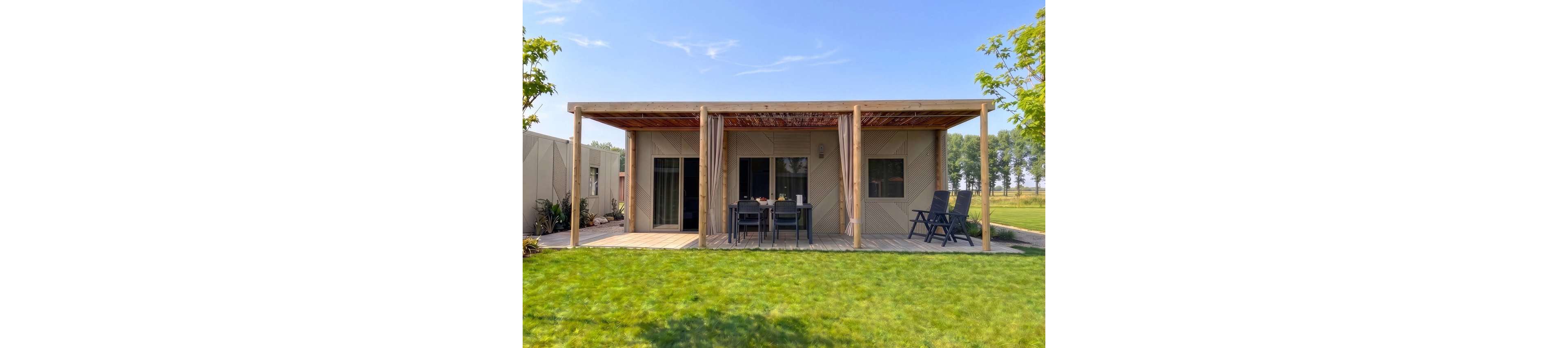 A modern single-story bungalow features a covered wooden patio opening onto a lush green lawn. Outdoor dining furniture is arranged at the center of the patio, with two lounge chairs positioned to the side. Large sliding glass doors and windows connect the interior to the outdoor space, allowing natural light to enter. The home is set in a peaceful rural environment with trees and open fields in the background under a clear blue sky.