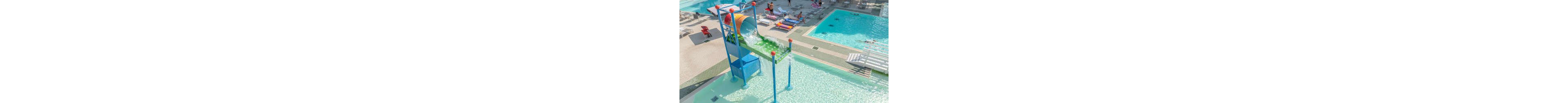 A large colorful tipping bucket pours water into a shallow splash pool at an outdoor resort. Nearby, people relax on lounge chairs and walk around the surrounding deck. Additional swimming pools and sunbeds are visible in the background, creating a family-friendly water park atmosphere. The scene is bright and sunny, emphasizing the clear blue water and playful structures.