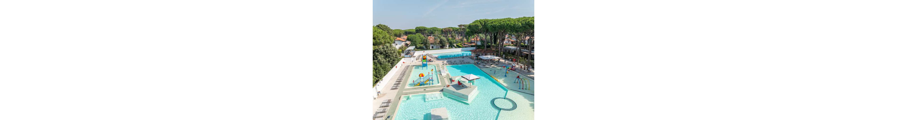 A large outdoor swimming pool complex features shallow pools and a colorful water playground for children. Sun loungers line the poolside, and a shaded seating area with umbrellas sits on a central platform. Additional pools and splash zones with water arches extend into the background. Tall pine trees and nearby resort buildings surround the aquatic area, creating a relaxed holiday atmosphere.