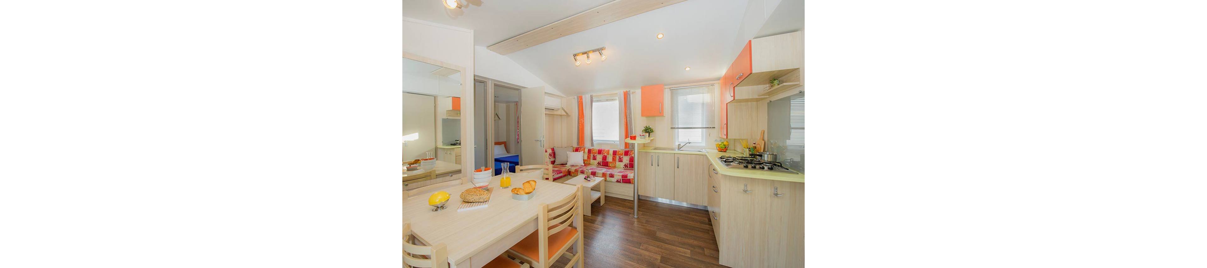 A small open-plan kitchen and dining area is shown in a bright, modern interior. Light wood cabinets and furniture contrast with orange accents on the cupboards and patterned sofa. A dining table is set with bread, fruit, and drinks, while the kitchen counter holds cookware and utensils near a gas stove. Large windows and a mirror reflect natural light, making the compact space feel airy and welcoming.