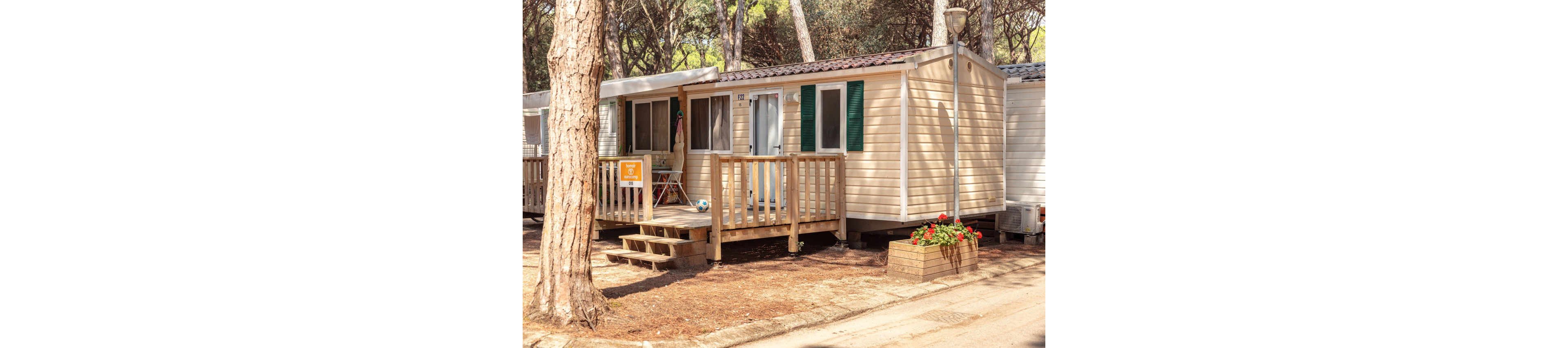 A beige mobile home cabin with a small wooden deck stands among tall pine trees in a forest campground. The deck features outdoor chairs, a table, and a colorful beach umbrella, suggesting a relaxed holiday setting. A soccer ball rests near the steps, adding a playful family vacation detail. A planter box with red flowers decorates the front, while neighboring cabins line the quiet dirt path.
