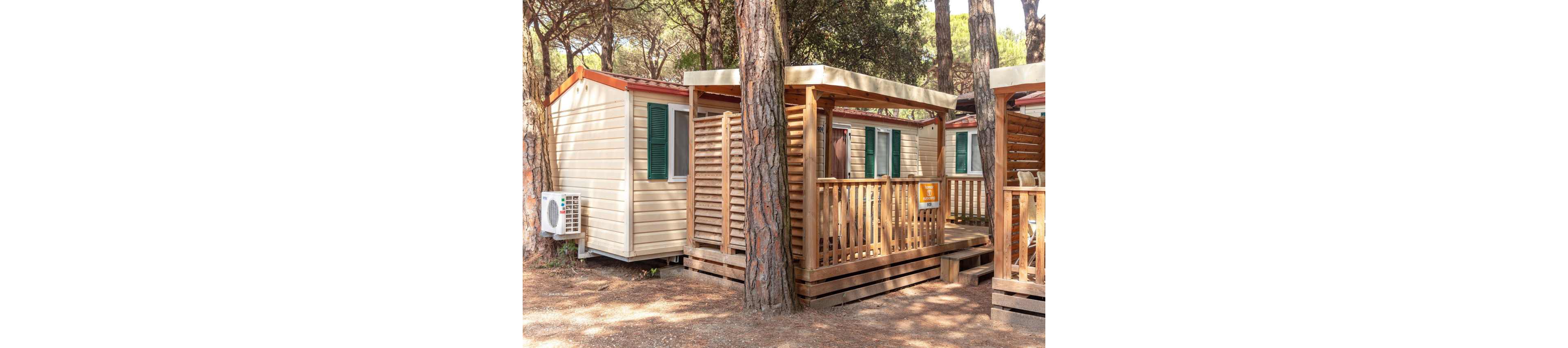 A compact mobile home with a wooden veranda stands among tall pine trees in a forest campsite. The cabin features cream-colored siding, green shutters, and a small covered terrace with railings. An outdoor air conditioning unit is mounted on the side, suggesting comfort in warm weather. Sunlight filters through the trees, creating a relaxed holiday atmosphere in a natural setting.