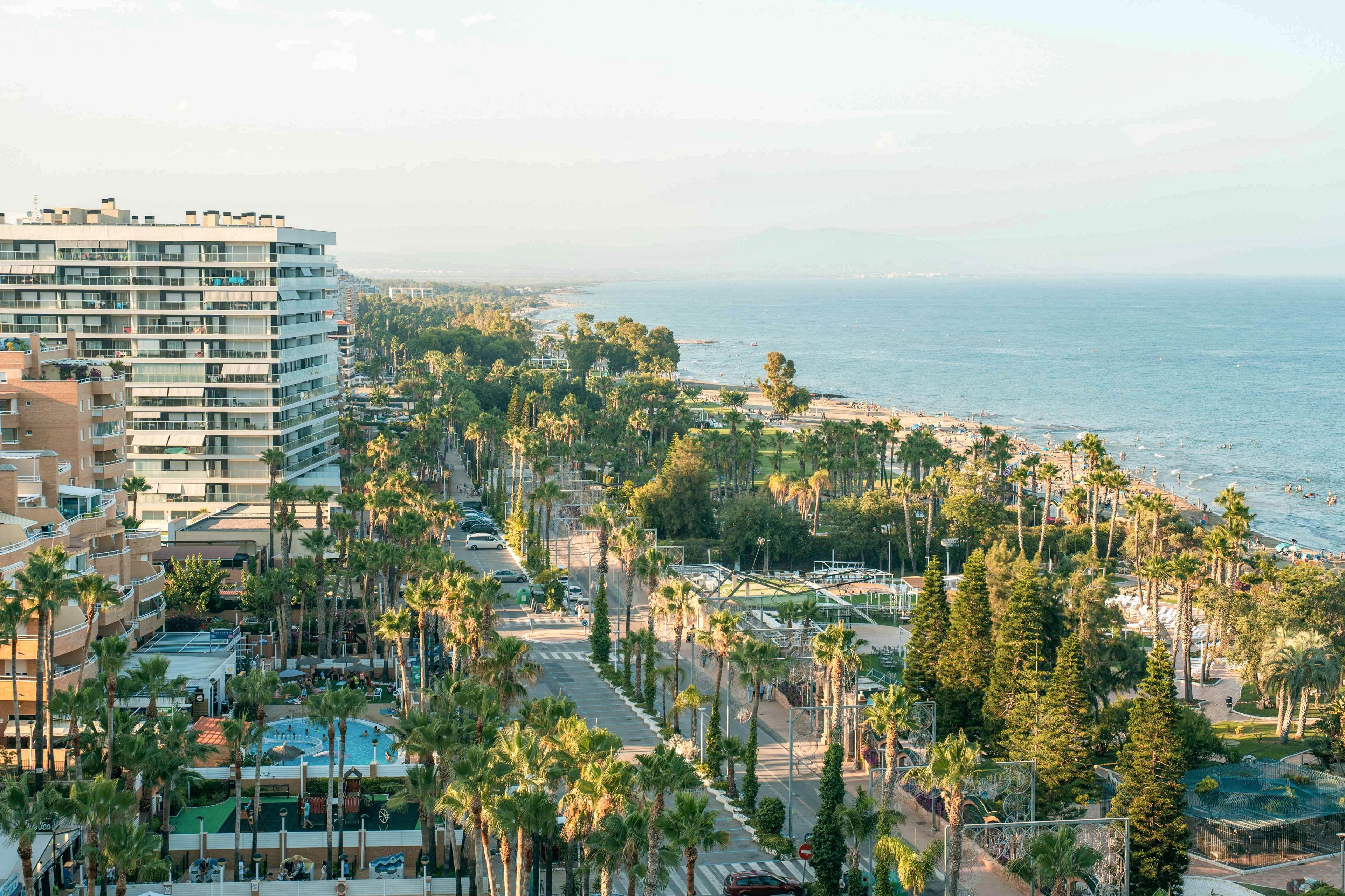 An elevated view captures a modern coastal city with mid-rise apartment buildings overlooking the sea. A wide avenue lined with tall palm trees runs parallel to the shoreline, leading the eye toward the distant horizon. The beach and waterfront promenade stretch along the right side, while pools and landscaped areas are visible among the buildings. Soft daylight and a light haze create a calm, relaxed seaside atmosphere.