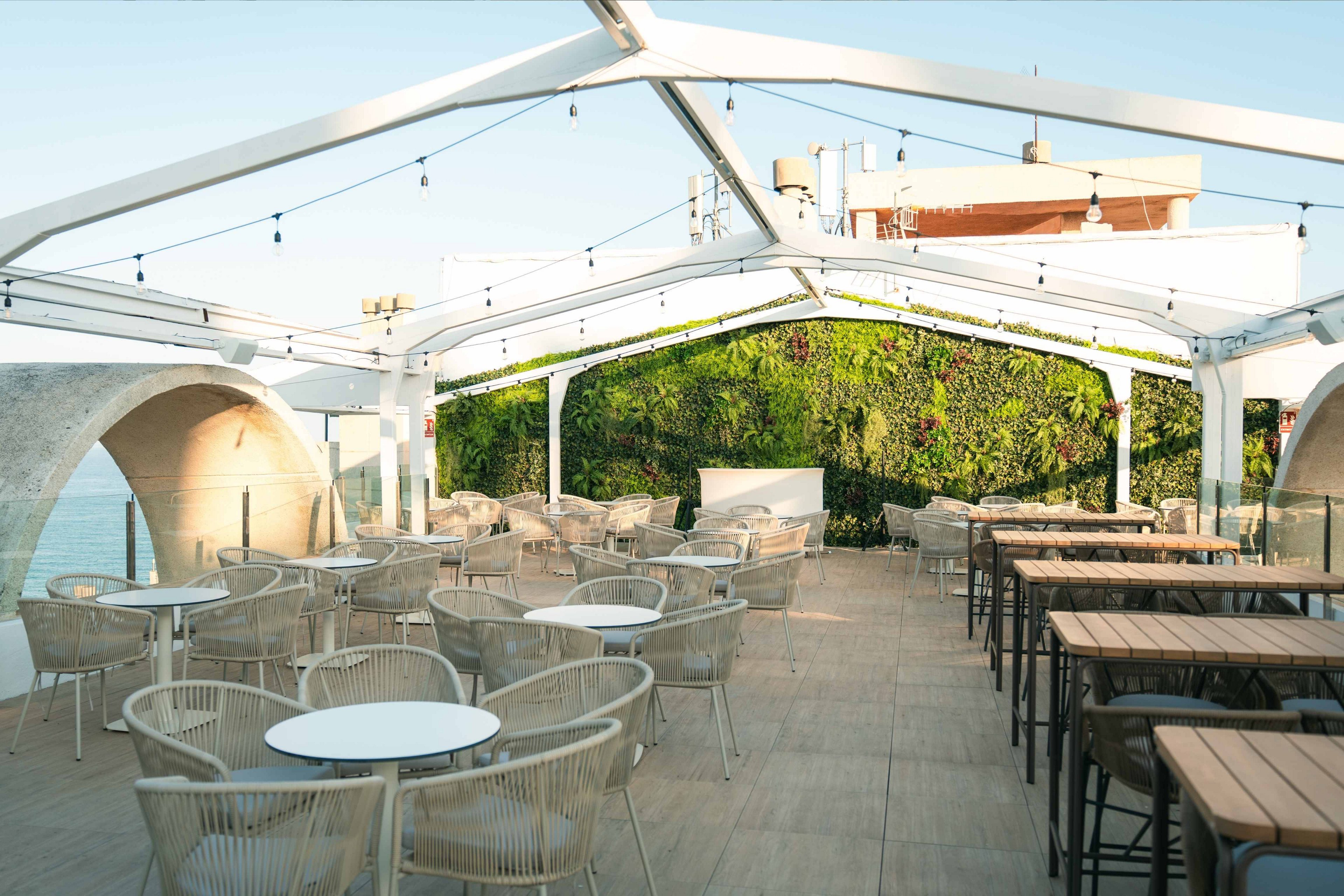An open-air rooftop terrace features rows of wicker chairs and wooden tables arranged neatly under a white metal pergola. String lights hang across the structure, suggesting a cozy atmosphere for evening gatherings. A lush green vertical garden covers the back wall, adding natural color to the bright, minimalist space. The setting overlooks a waterfront area, creating a relaxed and inviting environment for dining or social events.