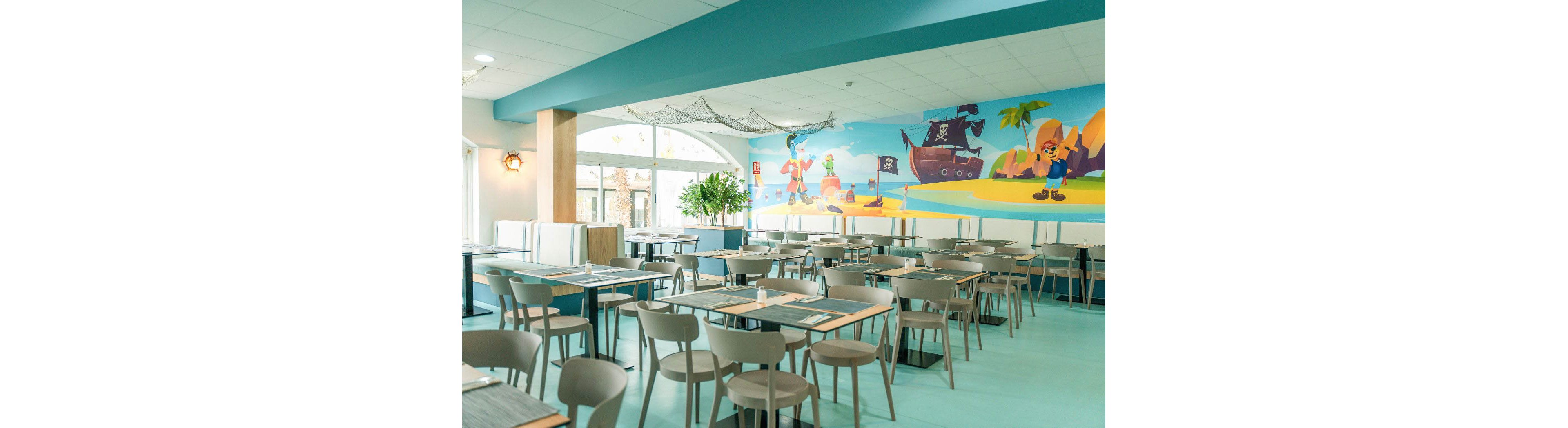 A light and airy restaurant dining area features rows of modern tables and chairs neatly arranged. Large windows let in natural daylight, illuminating the pastel blue floor and neutral furnishings. A colorful wall mural depicts a playful pirate scene with a ship, island, and cartoon characters, adding a child-friendly atmosphere. The space appears clean, spacious, and inviting for families and groups.