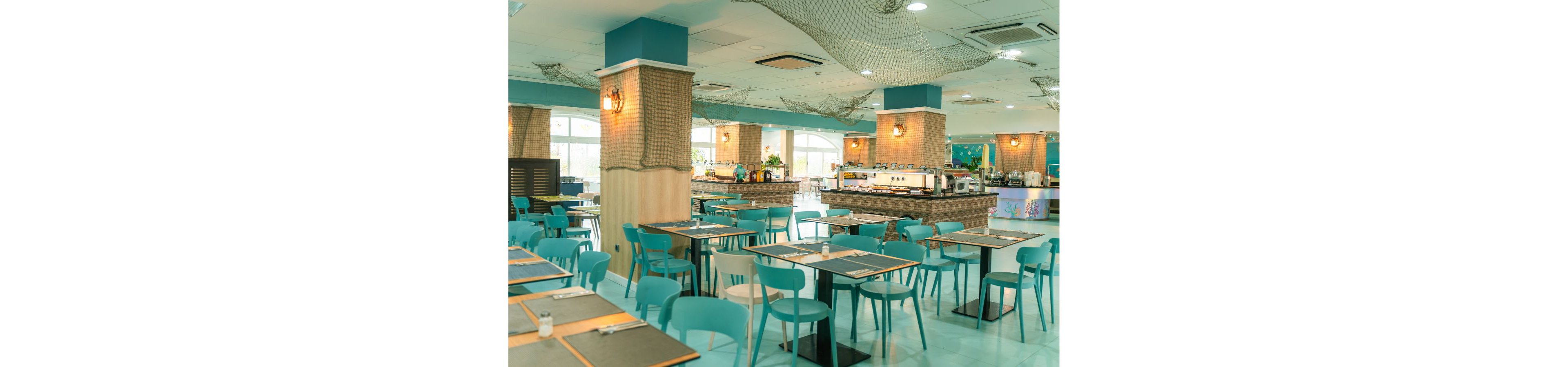 Spacious cafeteria interior featuring rows of turquoise chairs and dark tabletops. The setting appears clean and modern, with large windows letting in natural light. Decorative fishing nets and rope-wrapped columns create a coastal or nautical theme. A buffet or self-service counter is visible in the background, suggesting a casual dining environment.