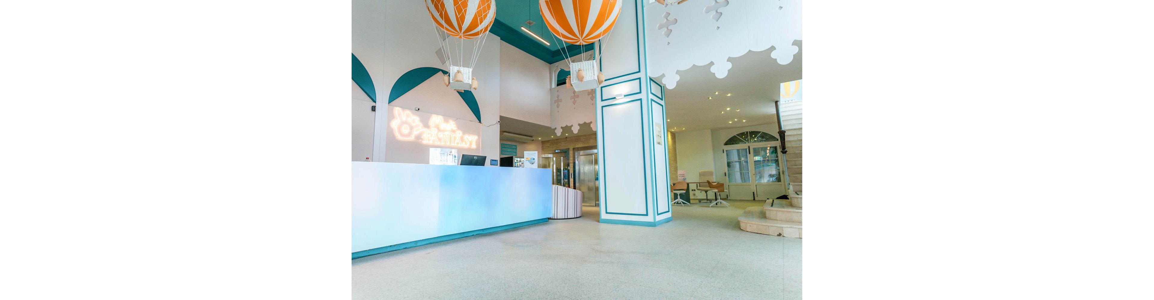 A family of four walks toward a reception desk in a bright, modern hotel lobby. The parents and two children are casually dressed in light, summery clothing as they approach the smiling receptionist. The interior features pastel colors, whimsical hot air balloon decorations, and soft ambient lighting. Large windows and open space create an airy, welcoming atmosphere suitable for vacation or resort stays.