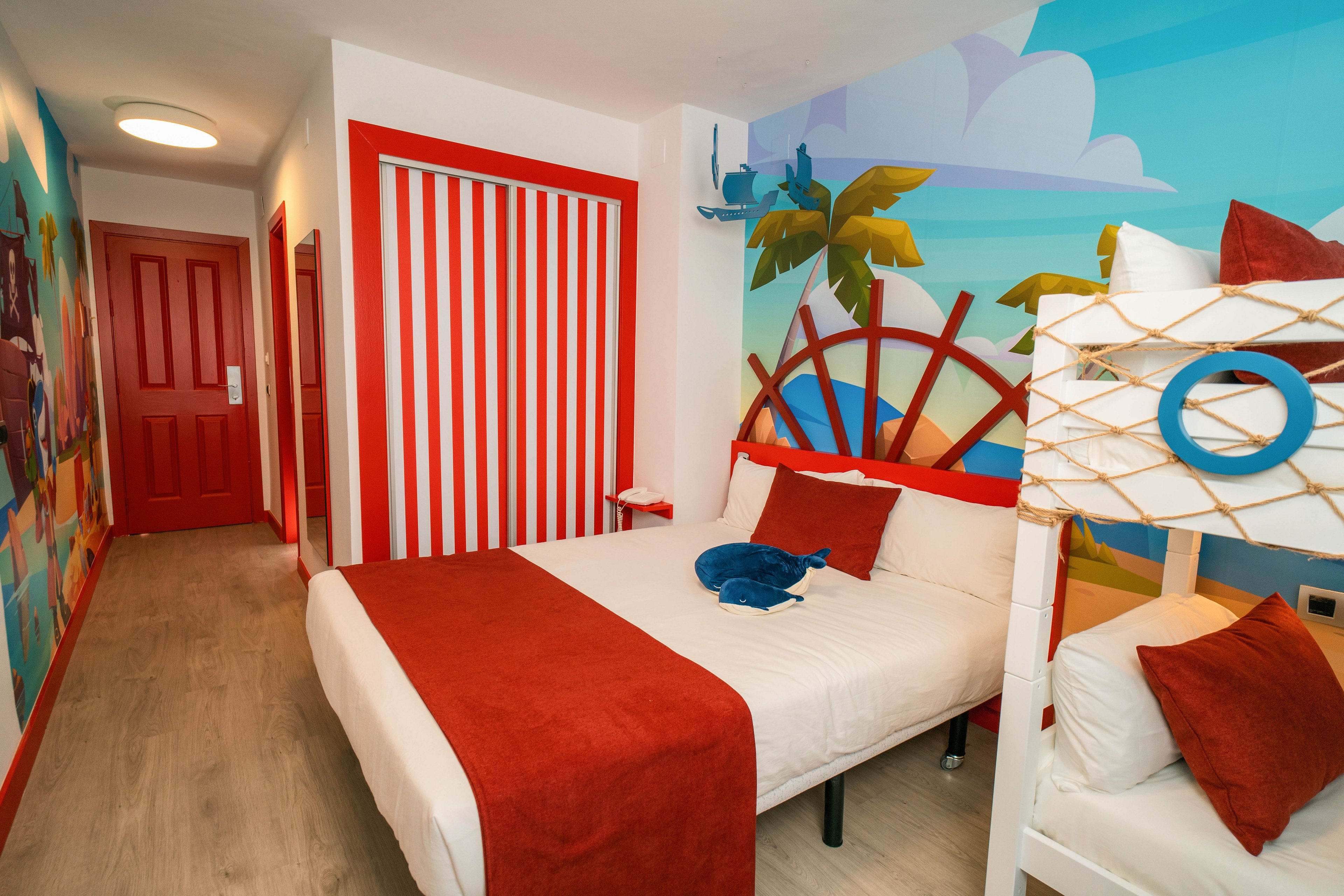 A bright family hotel room features a double bed with red and white bedding and a playful nautical theme. A bunk bed stands to the right, decorated with rope and circular blue accents. The walls are covered with colorful murals of palm trees, clouds, and ocean-inspired graphics, while a red door and striped closet add bold contrast. The overall design feels fun and inviting, ideal for children and family vacations.
