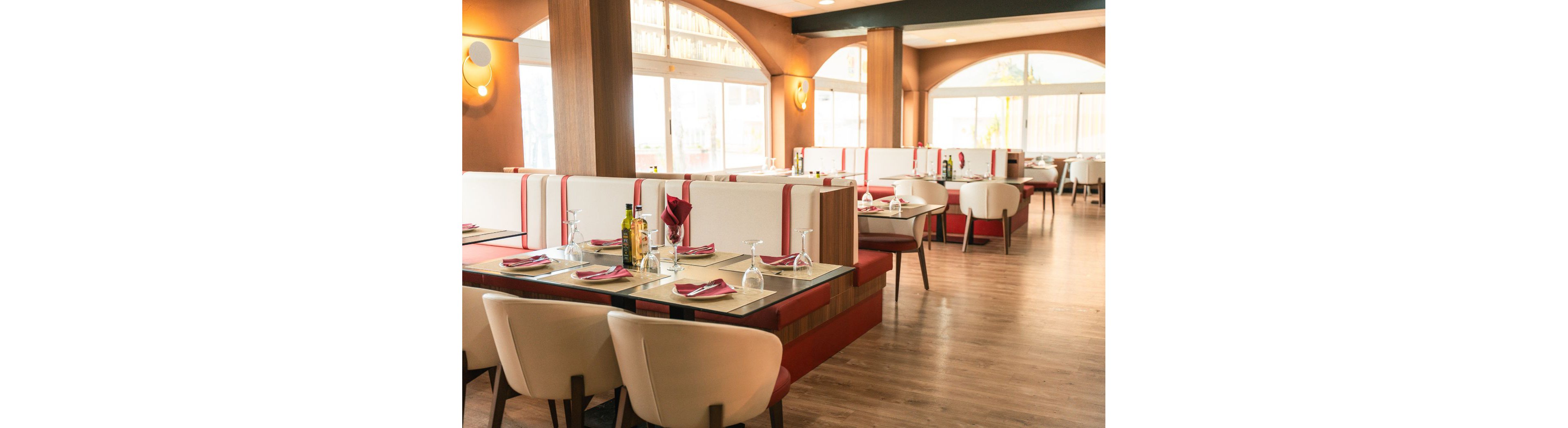 A contemporary restaurant dining area features neatly arranged tables and cushioned booths with red and white upholstery. Place settings with glasses, plates, and folded napkins are prepared on each table, along with a bottle of olive oil and condiments. Large windows allow natural light to brighten the warm-toned interior and wooden floor. The space appears clean, organized, and ready to welcome guests.